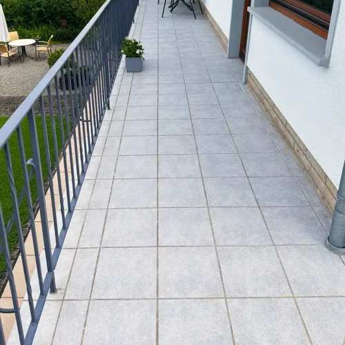 Gesamtansicht Balkon nach Balkonsanierung mit neuen hellgrauen quadratischen Fliesen, links Garten, rechts Hausfassade